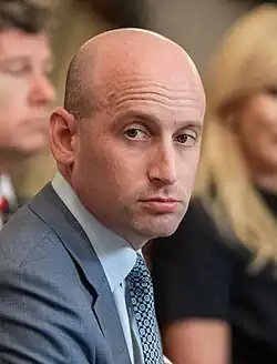 Miller facing frontward against a beige background giving a neutral expression.