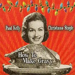 A sepia coloured image of a woman presenting a platter containing a large roast bird (possibly a turkey). Her smiling face is visible beyond the bird, her fingers appear below the platter. The background is red. A strand of coloured Christmas lights stretches across the top of the image. The artist's name is located to the left of the woman's head and the words, Christmas Single, are to her right. The title of the single is written over the image of the roast bird.