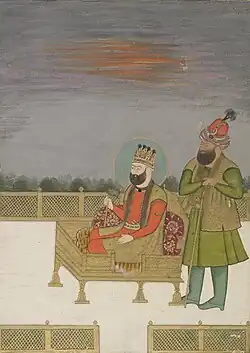 A color portrait of two men during sunset. One of them, a bearded man wearing a crown, is seated on the throne, while the other stands to his left