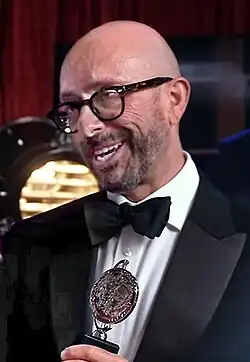 bald European caucasian man, approx 50 years old, with stubble and glasses, wearing black jacket, white shirt, black dickie bow tie, receives silver metal disc trophy