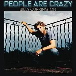 The cover has the artist wearing a blue t-shirt and jeans combination, crouching and having his right arm hold next to a black art deco fence. The song title is set above the image, colored in neon blue, with the artist's name below it in white.