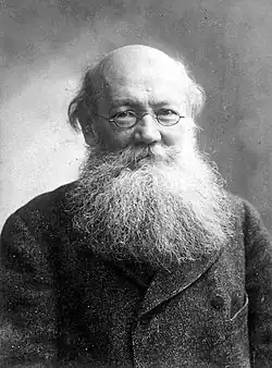 Bust portrait of an older man with no hair on his head, a wispy beard, and small glasses, in a coat