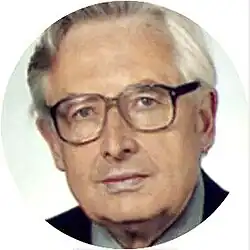 Colour portrait of an unsmiling, clean-shaven, middle-aged, white man with a square jaw, high brow, and greying hair combed back, wearing square glasses and a suit