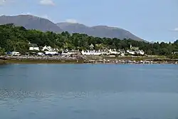 White painted cottages alongside a sea inlet, with grazing and trees behind taken from high up on a nearby crag, backed by a seascape having small islets in distance