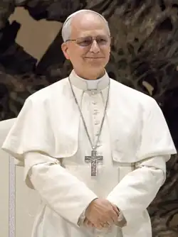 Photograph of Pope Leo XIV wearing papal regalia and slighty smiling. His dress consists of a white cassock with matching pellegrina and with white-fringed fascia, silver pectoral cross, and white zucchetto.