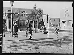 A group of approximately four Black children play on a swing set surrounded by buildings in 1938..