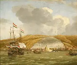 Two ships with sails and smaller boats with oars in a harbor, with a walled city and a citadel behind them and a steep hill in the background