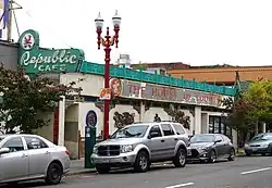 Photograph of the exterior of a building with a neon sign, parking pay station, and red lamp post; cars line the street.