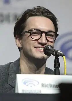 Richard Harmon wearing glassed, looking to the right of the camera and smiling.