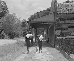 Two people in Rodney, Mississippi, walking down the street towards the Rodney Hotel and Pape store, barefoot with feed bags slung over their shoulders