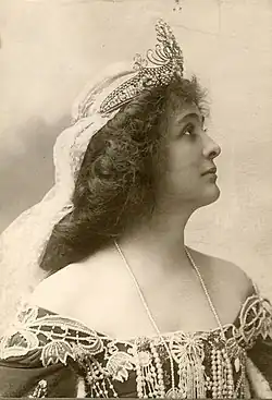 A white woman in profile, wearing a tiara and veil, with an off-the-shoulder gown trimmed in lace and pears, and a strand of pearls or beads.