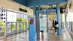 Renjong LRT platform with platform barriers with glass panels, a blue information stand with a clock, an emergency fire phone, and a map. A person with a backpack stands near the information stand, and residential buildings are visible in the background, with lush greenery between them and the platform.