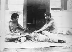Black-and-white photograph of two Samoan men tattooing a third. The third man is lying face down on the floor between the two men, one of whom is holding him down, while the other taps a stick into the man's back to create a tattoo.