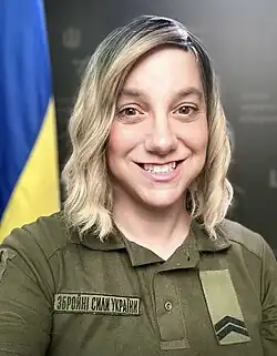 Portrait of Sarah Ashton-Cirillo in uniform, standing in front of an Ukrainian flag. She is a white woman with blonde hair, smiling at the viewer.