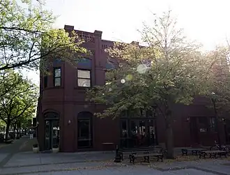 A two-story brick commercial building built in 1892. It has big windows and sits on the street corner, surrounded by wide sidewalks and trees.