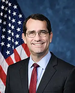 Official House portrait of Schmidt smiling in front of the U.S. flag, wearing a black suit, light blue shirt, and spotted red tie.