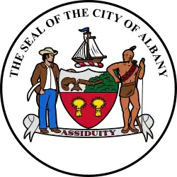 Circular seal with central images of a shield at center and sailing ship above it, with a European man to the left and a Native American to the right. The seal's edge reads "THE SEAL OF THE CITY OF ALBANY" with "ASSIDUITY" in a banner above the bottom.