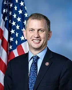 Official portrait of Casten from the 117th Congress. Sitting in front of an American flag, he wears a dark suit, a light blue shirt, and a blue tie.