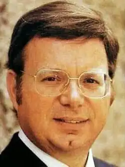 Colour portrait photo of of a smooth-shaven, half-smiling man with short brown hair, spectacles and sideburns.