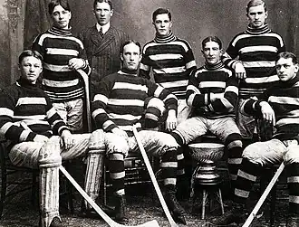 Seven men in hockey uniforms, four sitting in the front row, around the Stanley Cup trophy. Three stand in the back, with a man in a suit standing behind them.