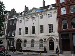 A four-storey, white bricked building, with two black doors. A flag pole is protruding from the centre of the building.