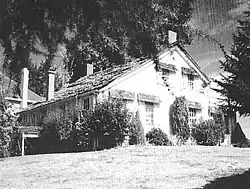 A modest two-story farmhouse with a peaked roof and four chimneys sits under a large tree at the top of a sloping lawn. Bushes and shrubs grow near the house on its two visible sides. A small part of a neighboring house, perhaps only 15 feet (4.6&nbsp;m) away, can be seen in the background.