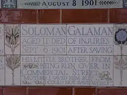 A tablet formed of five tiles of varying sizes, bordered by yellow and blue flowers in an art nouveau style. The tablet reads "Solomon Galaman, Aged 11 died of injuries, Sept 6 1901 after saving his little brother from being run over in Commercial Street, 'Mother I saved him but I could not save myself.'"