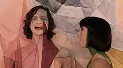 Upper body shot of a man (left) and a woman turned towards him. The man is taller and has dark stragly hair. The woman has dark hair cut in a bob. Both have skin painted in coloured geometric designs. Both are singing. The backdrop has a similarly coloured design closely matching the design on the two people.