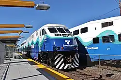 The front of a Sounder locomotive is seen on a track that passes through a station with a wide, uncovered platform. Another Sounder train's passenger car is seen behind the locomotive.