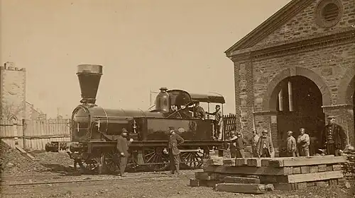 A 2-4-0 locomotive at an engine shed with workers standing by
