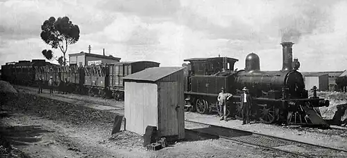 South Australian Railways broad-gauge K class steam locomotive with a water tank car, 5 open wagons, 2 passenger cars and a brakevan behind. Two crew members are posed by the locomotive. A weighbridge is in the foreground.