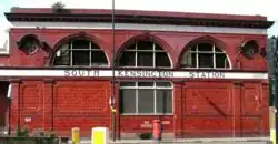 A two-storey red-glazed building with a single rectangular window on the lower floor and three large semi-circular windows on the upper floor flanked by two small circular windows. A white band between the floor levels displays "South Kensington station".