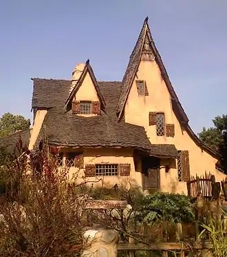 A whimsical storybook-style house with an asymmetrical pointed roof, small crooked windows, and rustic stucco walls, surrounded by an overgrown garden