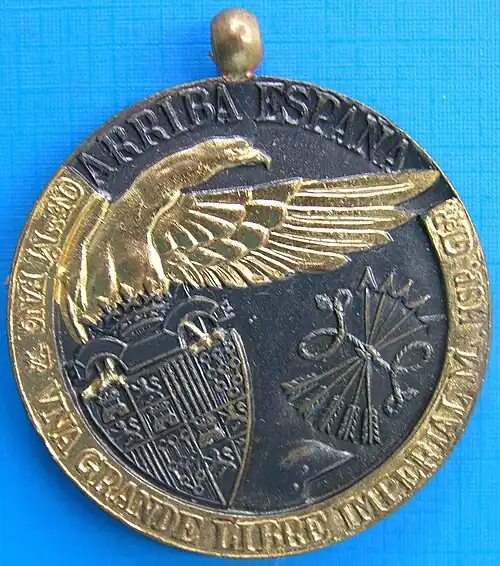 Photograph of a bimetallic medal. On a dark background metal, a coat of arms including quarters with a castle, a lion, four vertical bars and the chains of Navarre. The coat of arms is topped by a yoke and a crown. Also on the background, a military helmet and four arrows tied with a string. The top of the coat of arms is held by a golden eagle looking to the right and extending its wing to the right. Surrounding the medal, in the dark metal "ARRIBA ESPAÑA". In the golden metal, "[Illegible] M̊ [illegible] [a victor symbol] VNA GRANDE LIBRE IMPERIAL M. HSP. GLOR.