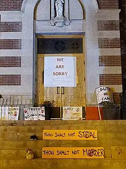 A church sign saying "We are sorry" hangs on the doors of the co-cathedral. The sign has messages scribbled on it including "Sorry does not cut it" The steps leading to the Cathedral are covered in signs from an earlier protest. The signs read: "The church is not the victim.", "Truth before reconciliation.", "You can't wash us away.", "Duty of care.", "Thou shalt not steal." and "Thou shalt not murder."