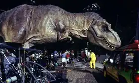 A life-sized Tyrannosaurus robotic model, with hydraulics where the dinosaur's feet would be, touches a car in a movie set.
