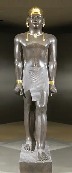 Statue of Taharqa. His name appears on the center of his belt: (tꜣ-h-rw-q, "Taharqa"). The statue is 2.7 meters tall. Taharqa has a striding pose, the arms held tight, and holds the mekes staff. He wears a shendyt or pleated kilt and on his head is a double-uraeus skullcap, possibly signifying his rule over Nubia and Egypt.[1] (Louvre Museum, color reconstruction of the jewelry through pigment analysis).[2]