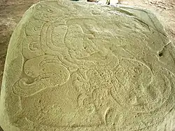 a flat stone with a sculpted bas-relief design. Two clawed feet at the left support a cartouche containing a seated figure, seen from the side with head to the right.