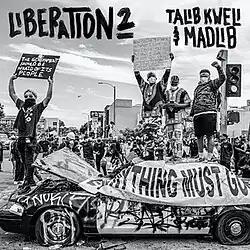A black and white photo of a crowd in street wear and masks, standing on top of a vandalized car, holding up signs
