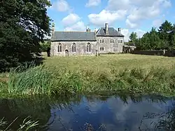 Seen beyond a river and a field is a simple stone chapel with three arched windows and a door; to the right is a two-storeyed stone house
