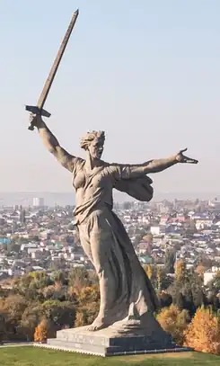 A massive sculpture of a woman with an outstretched arm holding a sword. The statue is dynamic, with the woman's robes appearing to billow in the wind, adding a sense of movement and urgency. Her other arm is extended forward, as if beckoning or calling out.