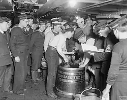 A group of men in World War I uniforms of the Royal Marines and Royal Navy, some of whom are holding metal pails, standing adjacent to a wooden tub inscribed with "God Save the King". A man scoops liquid from the tub (the rum ration) and pours it into the metal pails, as a Petty Officer looks on from a logbook. They are standing belowdecks on a ship with a low ceiling, with pipes and cables snaking overhead.