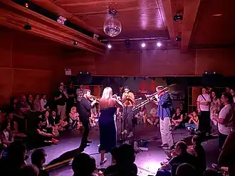 Two trumpeters and two trombonists perform in a circle surrounded by audience members in a wood-paneled room