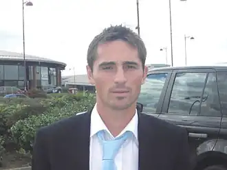 A head and shoulders of a man, wearing a black blazer, white shirt and blue tie.