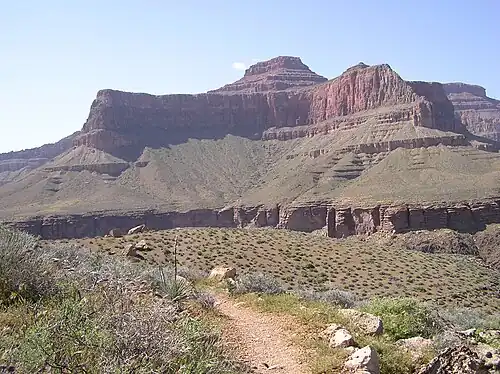 View of Tower of Set peak and sub-unit cliff section from Tonto Trail, Granite Gorge, north of Mohave Point, Grand Canyon Village, South Rim. The peak is behind and separated from a cliff unit (with small prominence), in front-(photo center, right, Tower of Set (peak) to its left). Vertical erosion in cliff of Redwall Limestone, upon horizontal Muav Limestone cliff.[19] The Tapeats Sandstone sits in foreground on Granite Gorge, and is seen as thinly-bedded. The slope-former above is the (dull-greenish)-Bright Angel Shale with thin, inter-bedding, as well as one resistant cliff unit. The Redwall Limestone cliff section in Grand Canyon is about 450 feet (137&nbsp;m) thick.[20]