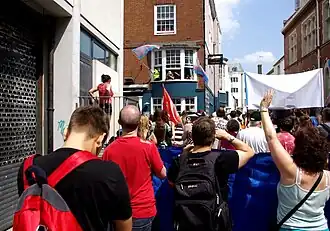 A group of people marching, photo taken from behind. Several trans pride flags are being waved.