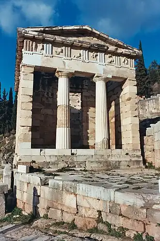 Color photograph of the front of a Classical Greek building located in a lightly forested, hilly landscape.