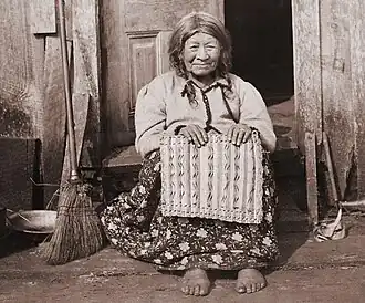 An elderly Clatsop woman sitting in front of a wooden door, wearing western style clothes. She is holding a traditional basket.