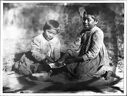 Two Havasupai children, the daughters of Chickapanagie's, enjoying a melon, ca. 1900. Both are wearing checkered dresses and are barefooted. They sit on a blanket with a zig-zag pattern with their hands in a half melon between them. Their hair is cut very short.