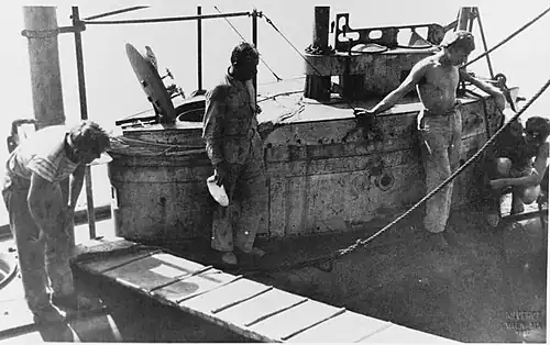 The conning tower of a submarine while docked. Four men stand around the conning tower while a gangway plank has been laid down next to it. The open hatch of the submarine and various ropes mooring the ship can also be seen.